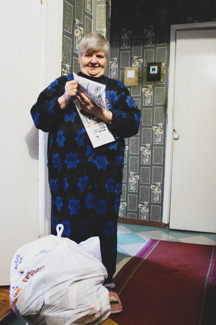 Elderly woman in Berdyansk, Ukraine wearing a blue dress standing in her apartment while holding a bag of FMSC food