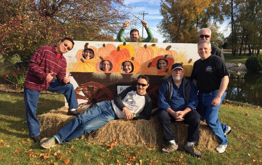 Group of adults on a hay bale in fall attire smiling for a photo, some with heads through pumpkin sign in Minnesota, USA