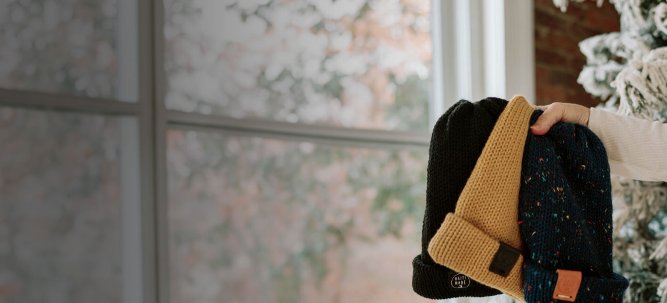 A hand holding 3 FMSC MarketPlace beanies beside a scenic window