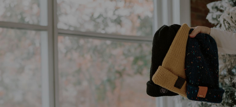 A hand holding 3 FMSC MarketPlace beanies beside a scenic window