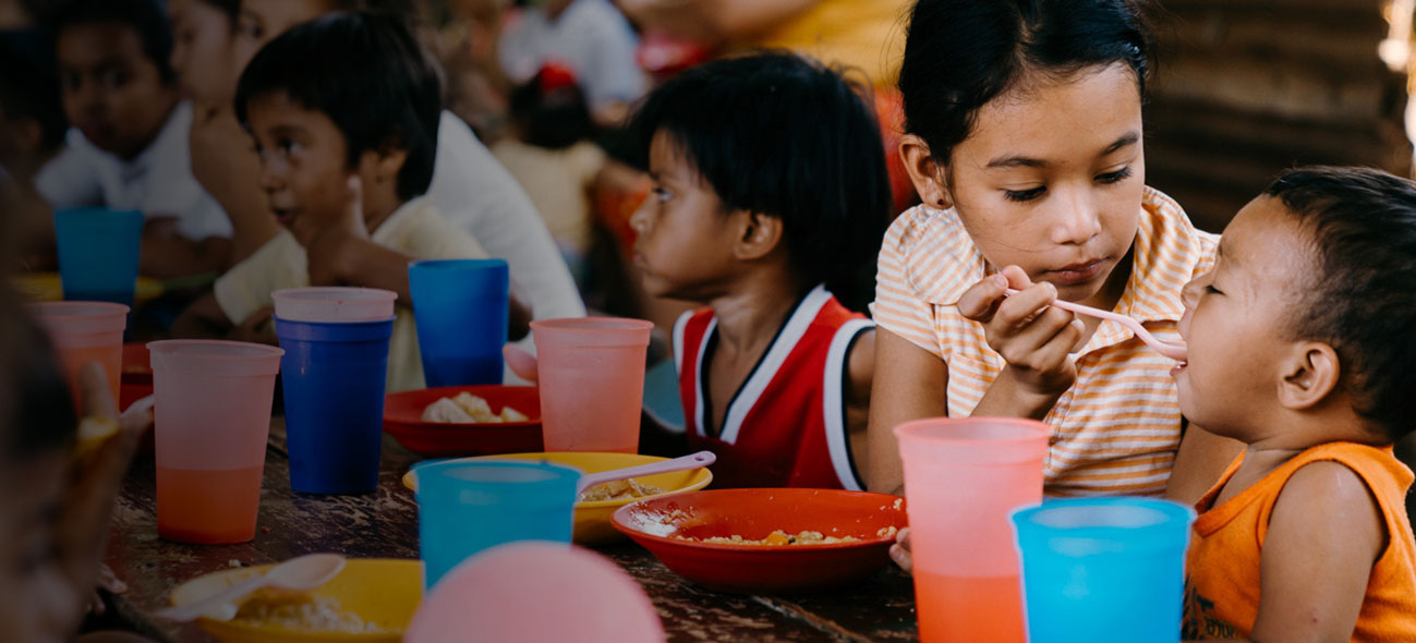 Children sitting at a table eating FMSC food, where a girl is spoonfeeding some to a toddler.