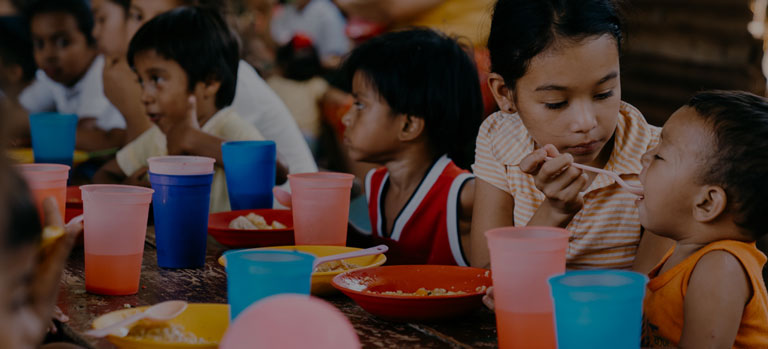 Children sitting at a table eating FMSC food, where a girl is spoonfeeding some to a toddler.